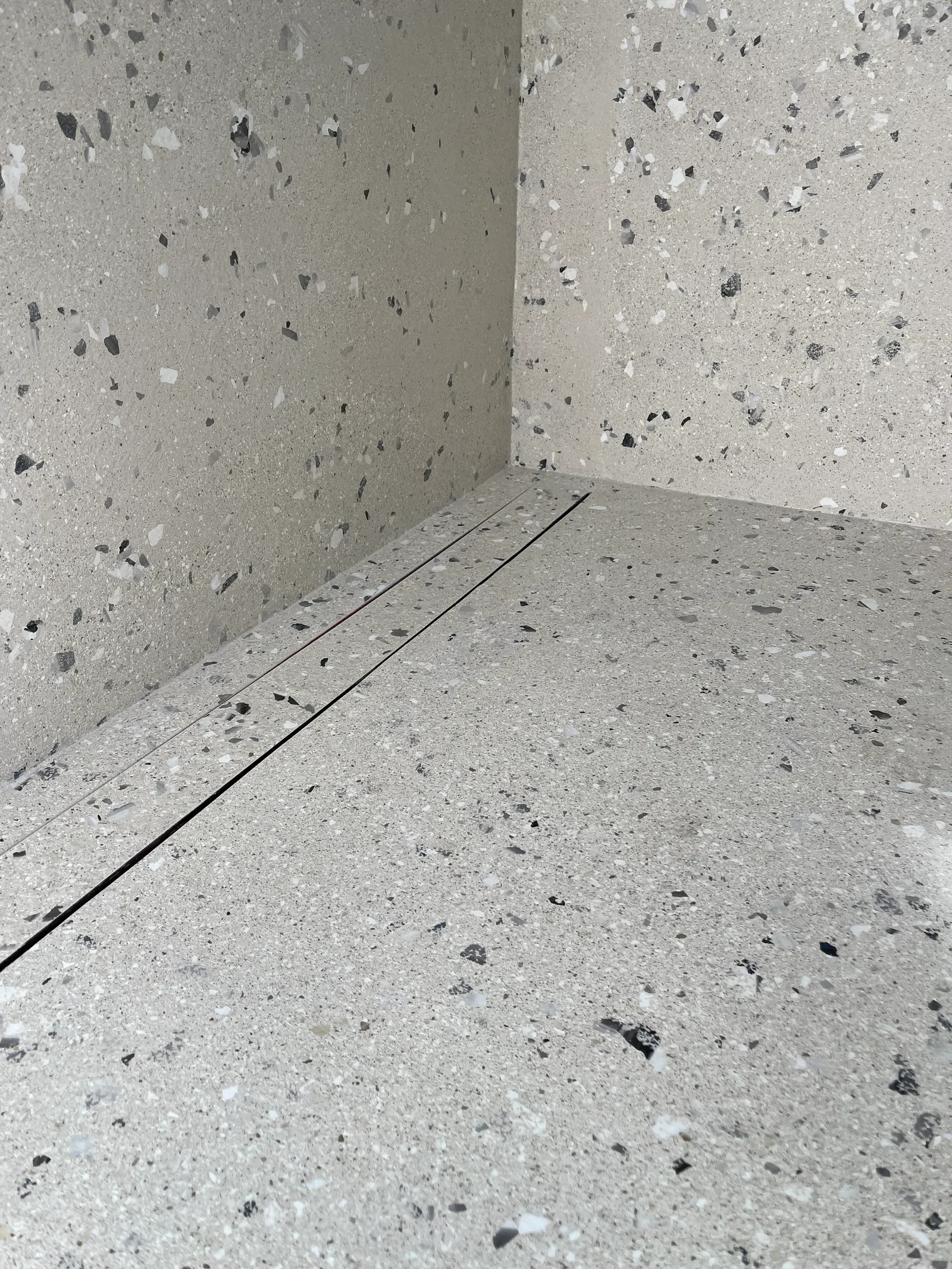 Een smalle lineaire afvoer langs de wand in een grijs gespikkelde douchevloer.